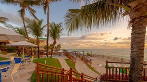 Un resort frente a la playa con mobiliario exterior, palmeras y una escalera de madera que da a la zona de la playa.