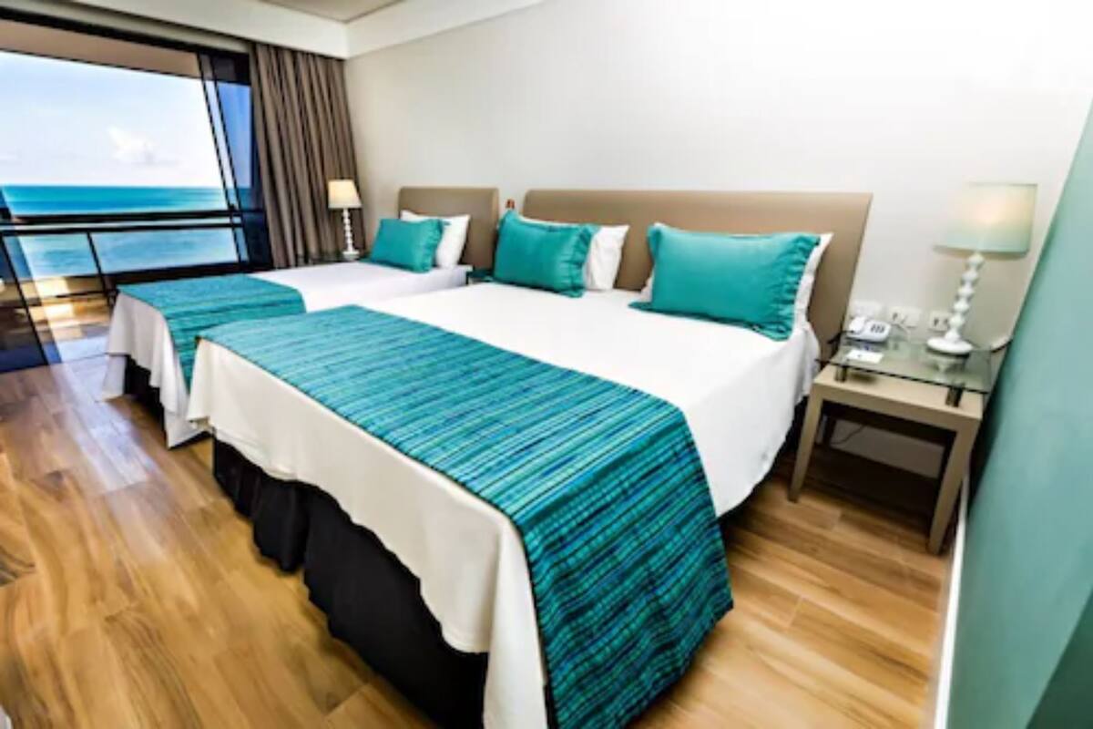 A hotel room with two beds, a large window with a view of the ocean, a wooden floor, and a nightstand with a lamp.
