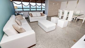 Uma sala de estar moderna com um sofá branco, um banquinho e um espaço de refeições com cadeiras brancas e uma mesa de vidro.