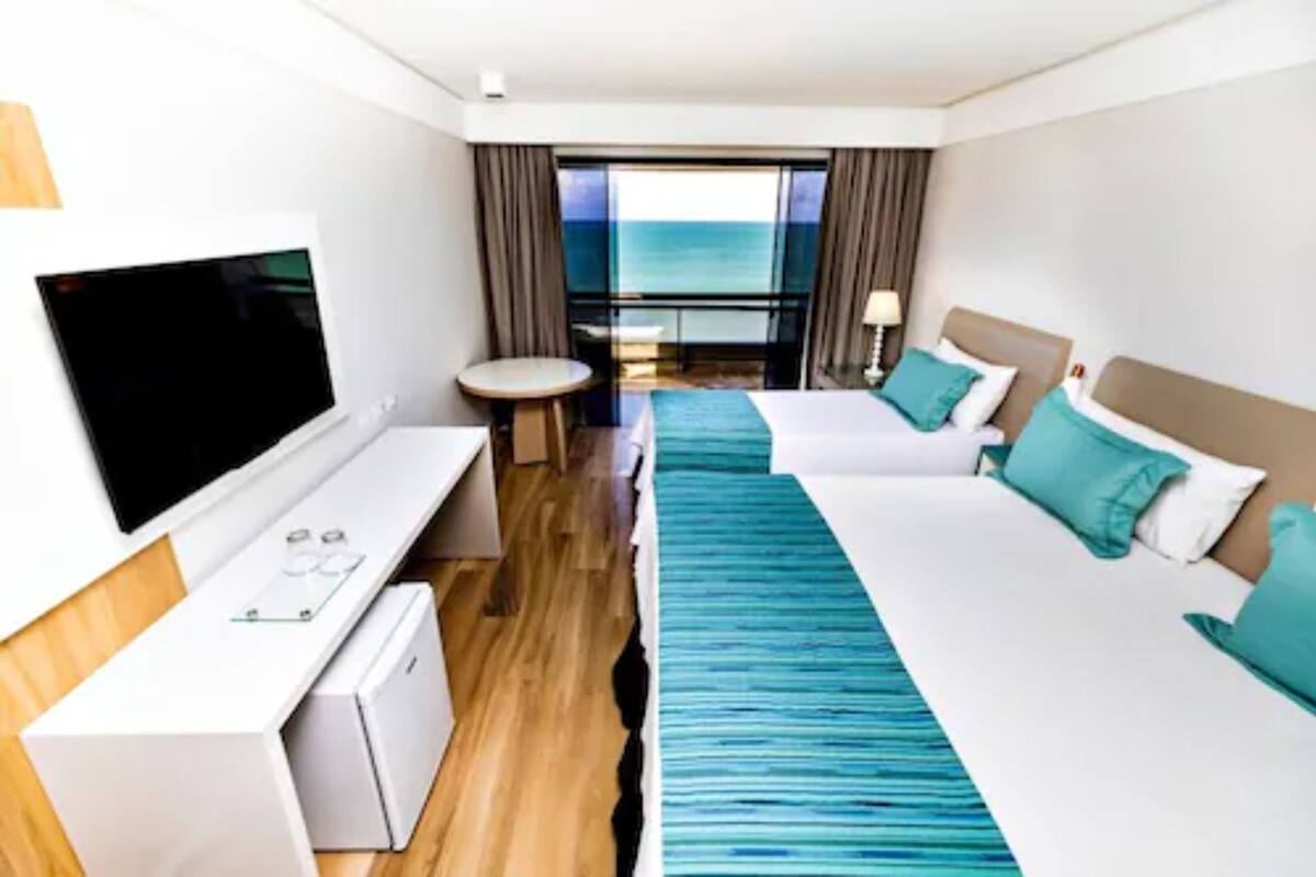 A hotel room with a bed, a desk with a TV, a small table, and a view of the ocean.