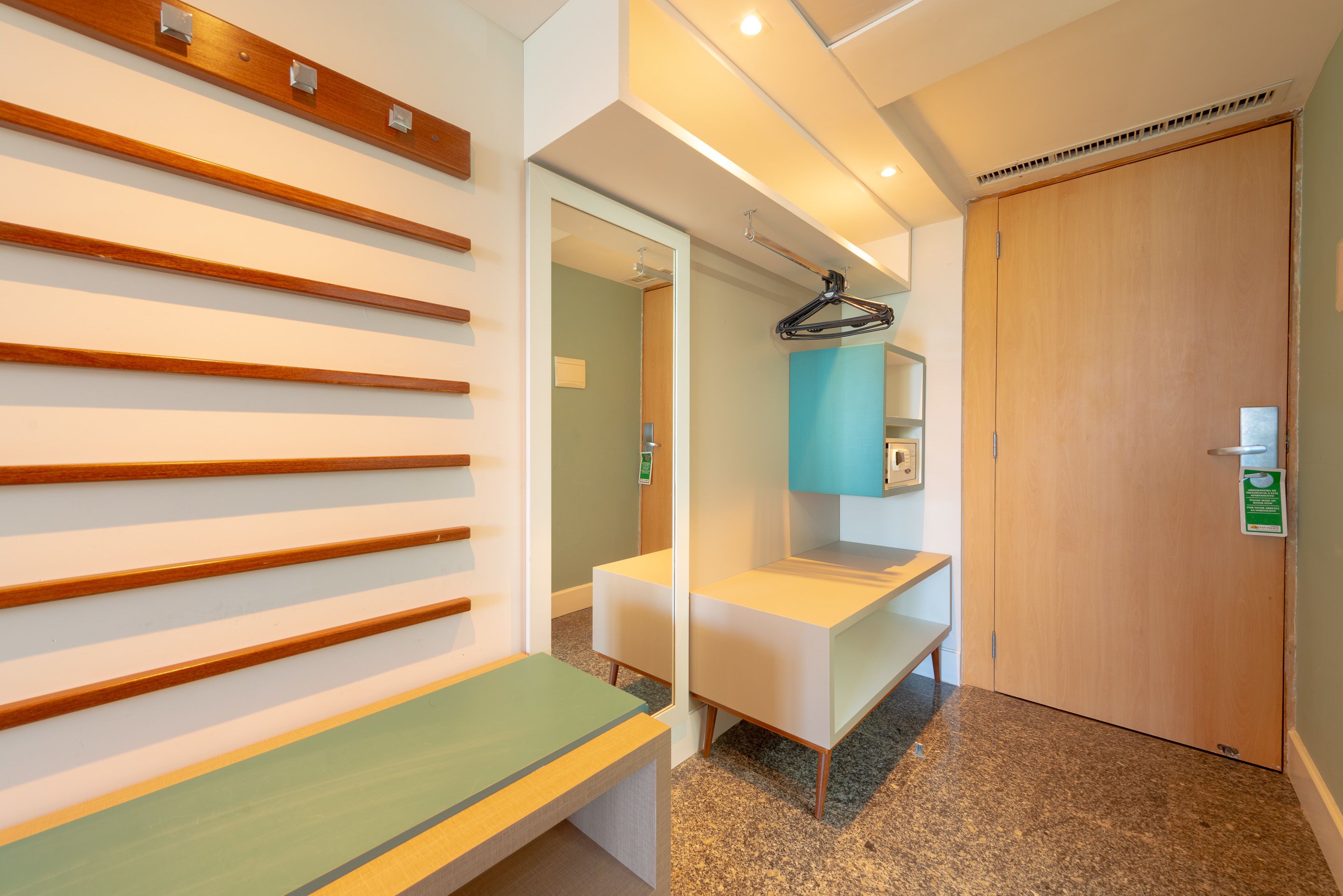 A modern hotel room with a wooden slat wall, a bench, a mirror, and a door.