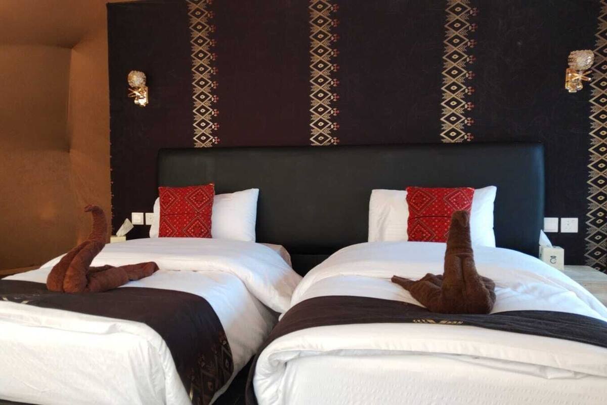 A hotel room with two beds, each with white linens and brown throws. There are decorative pillows and a dark headboard with a patterned design.