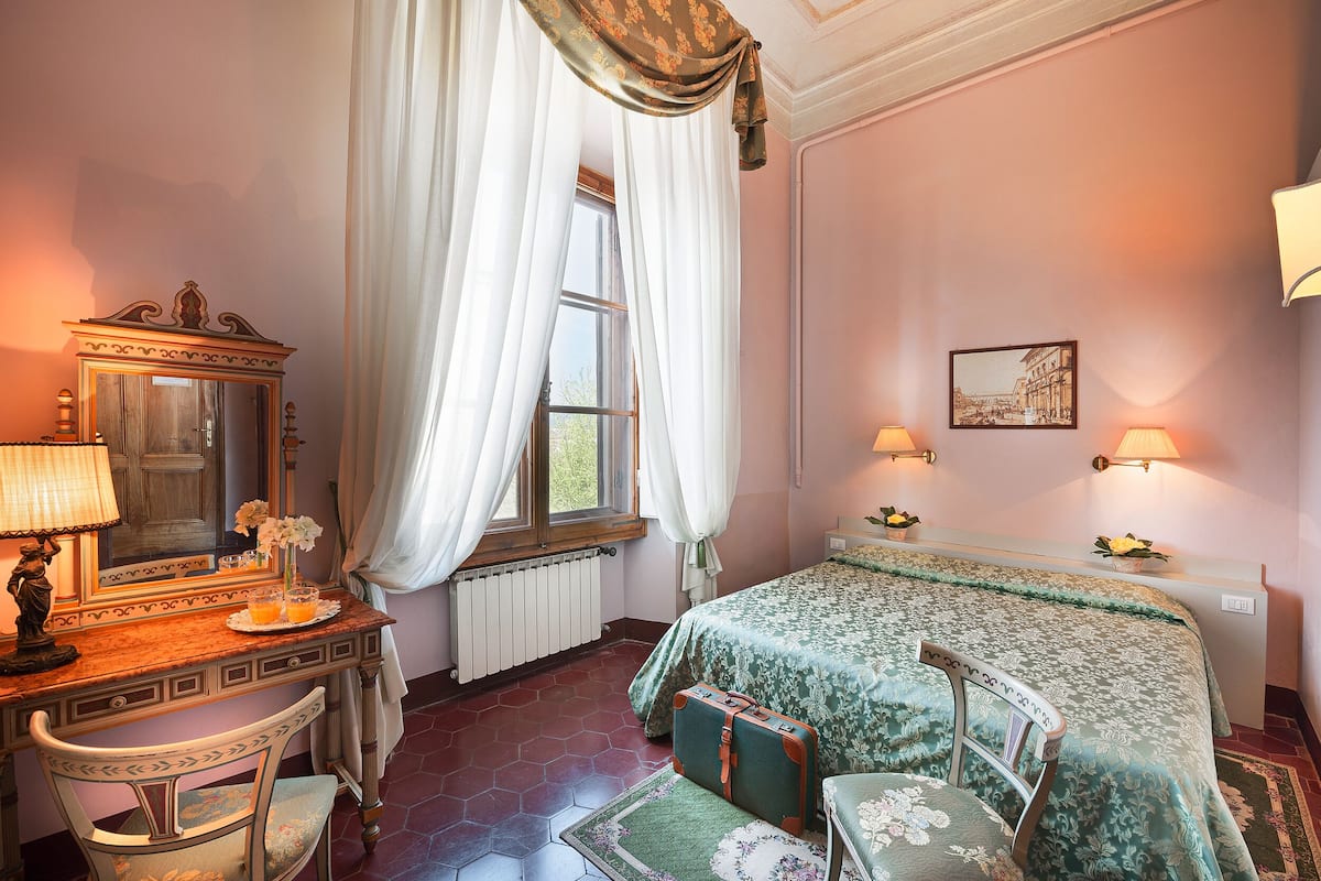 Une chambre à coucher comprenant un lit, un bureau avec un miroir, une chaise, une fenêtre avec des rideaux, un radiateur et des lampes fixées au mur.