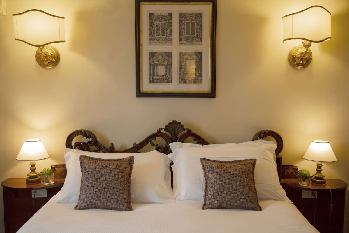 Un lit bien fait, avec du linge de lit blanc et des coussins à motifs, flanqué de deux tables de chevet sur lesquelles reposent des lampes et de petits objets décoratifs. Un tableau encadré est accroché au-dessus du lit.