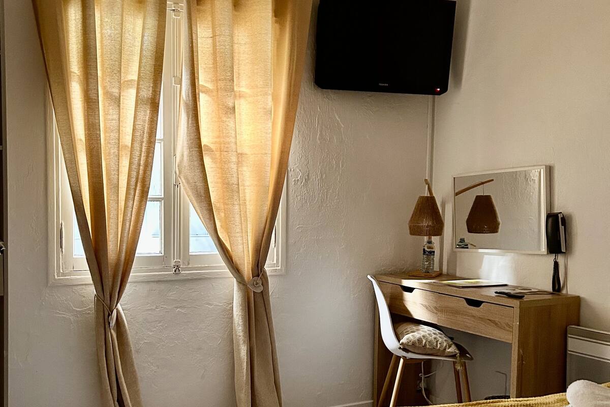 Un dormitorio con cama, escritorio con silla, televisor montado en la pared y ventana con cortinas.