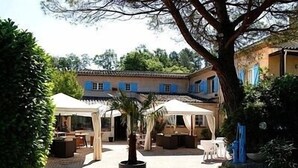 A courtyard with outdoor seating, a large potted plant, and a building with blue shutters in the background. - La Bastide des Golfs (Grasse)