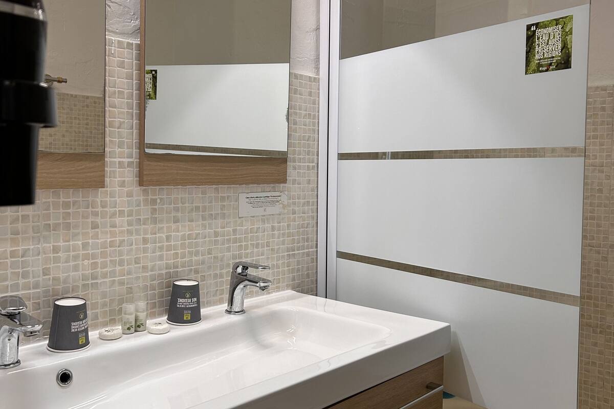 Un baño moderno con un lavabo blanco, dos grifos y un espejo encima. Debajo del lavabo hay armarios de madera y un perchero a la derecha.