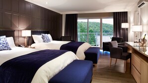 Habitación de hotel con dos camas, un escritorio, una silla y un balcón con vistas a zonas verdes.