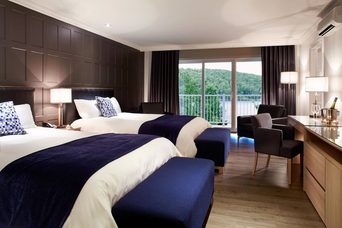 A hotel room with two beds, a desk, a chair, and a balcony with a view of greenery.
