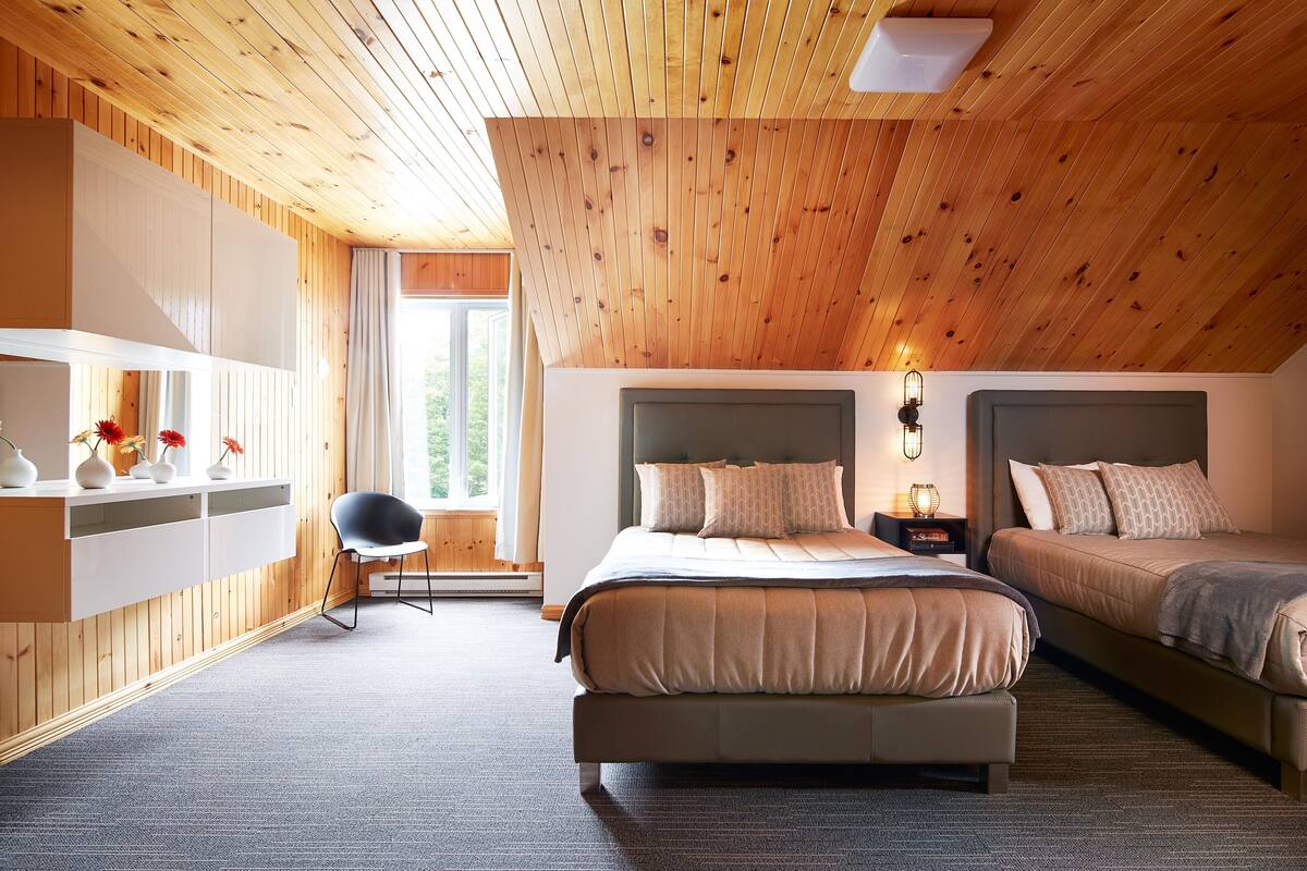A bedroom with two beds, a wooden ceiling, a built-in wardrobe, and a window with a view of greenery.