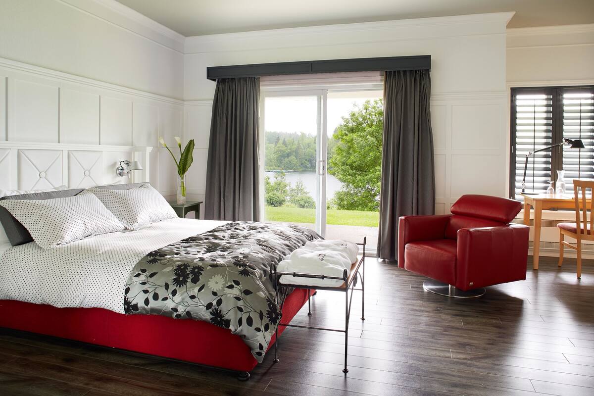 A bedroom with a bed, a red chair, a table, and a view of a lake.
