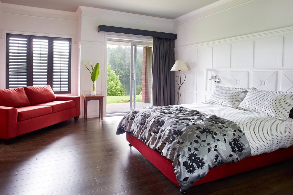 A bedroom with a red sofa, a large bed with a floral bedspread, a nightstand, and a view of greenery through the window.