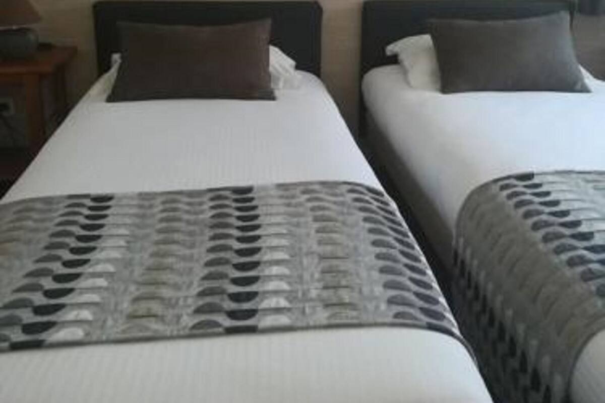 A hotel room with two beds, each with a pillow and a blanket. There is a nightstand with a lamp and a radio. The walls are tiled.