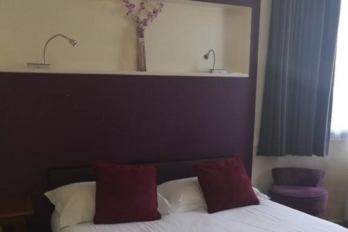 A hotel room with a bed, pillows, a red blanket, a vase with flowers, and a nightstand.