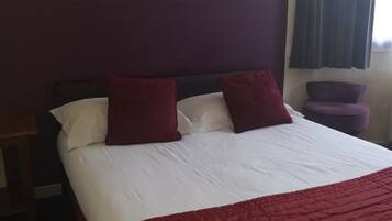 A hotel room with a bed, pillows, a red blanket, a vase with flowers, and a nightstand.