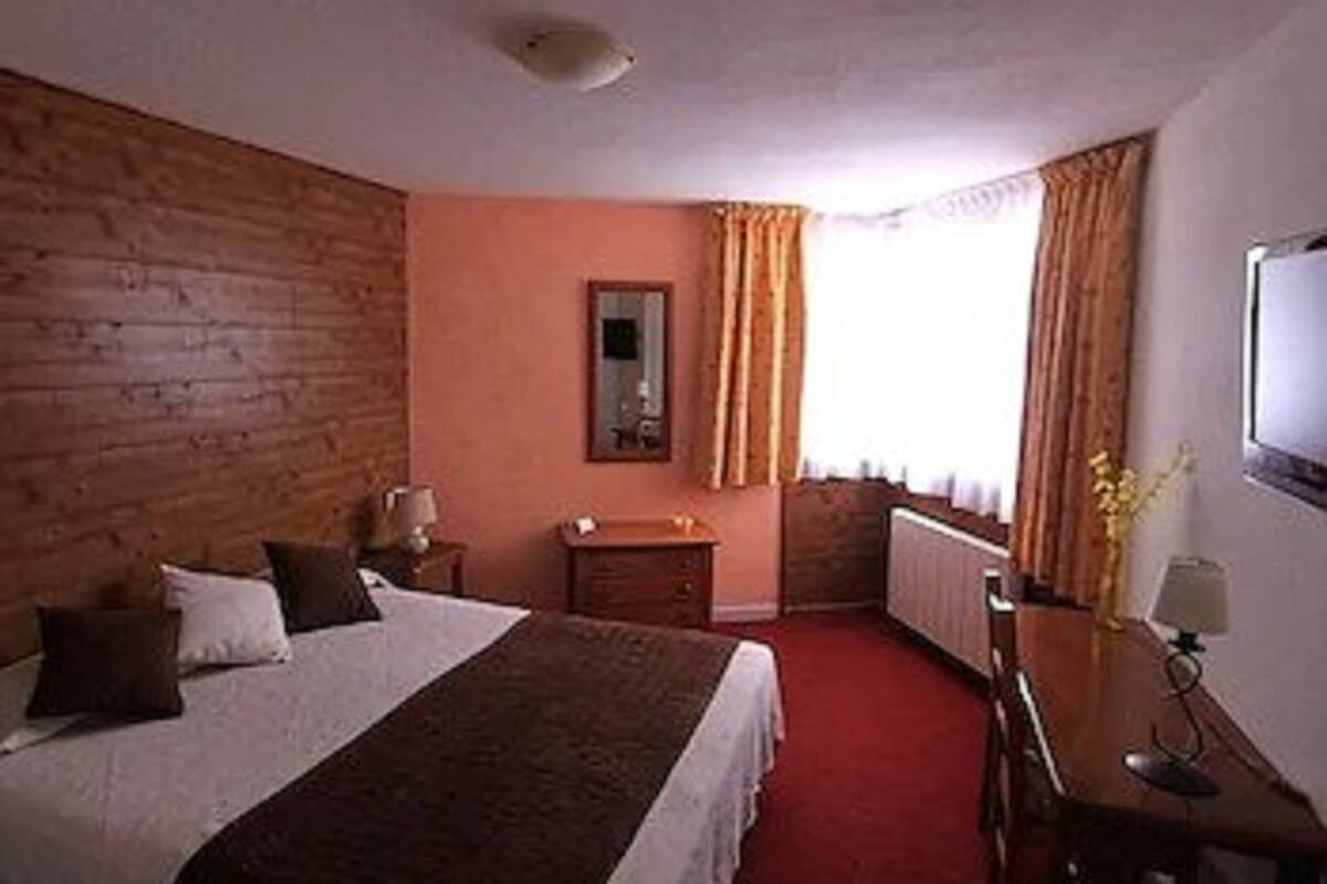 A hotel room with a wooden headboard, a single bed, a nightstand, a dresser, a mirror, and a window with curtains.
