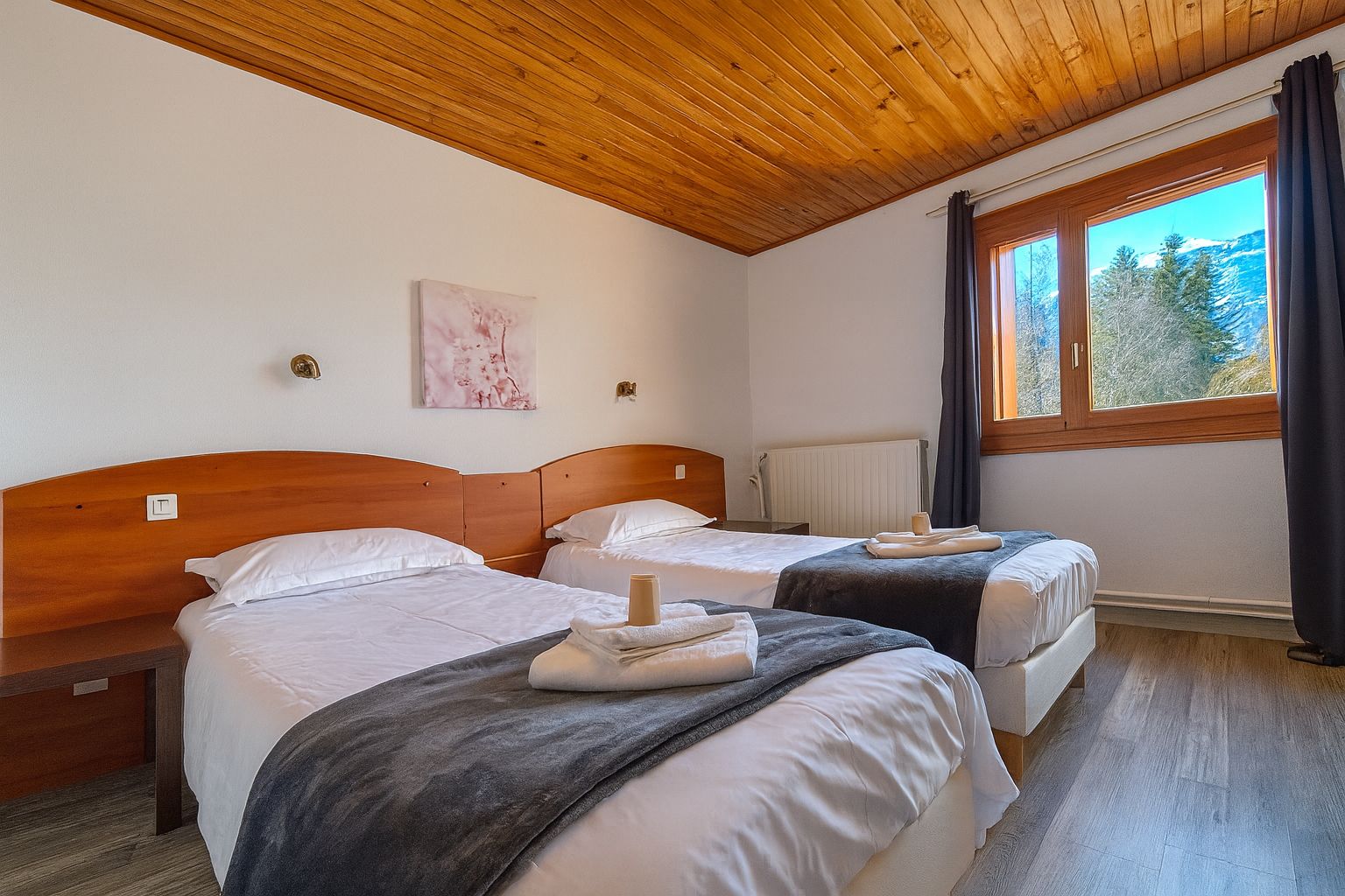 Een kamer met twee bedden, houten hoofdeinden, een raam met uitzicht op bomen en een radiator.