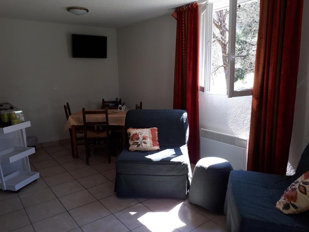 Ein Zimmer mit Esstisch, zwei Stühlen, einer Couch, einem an der Wand montierten Fernseher und einem Fenster mit Vorhängen.