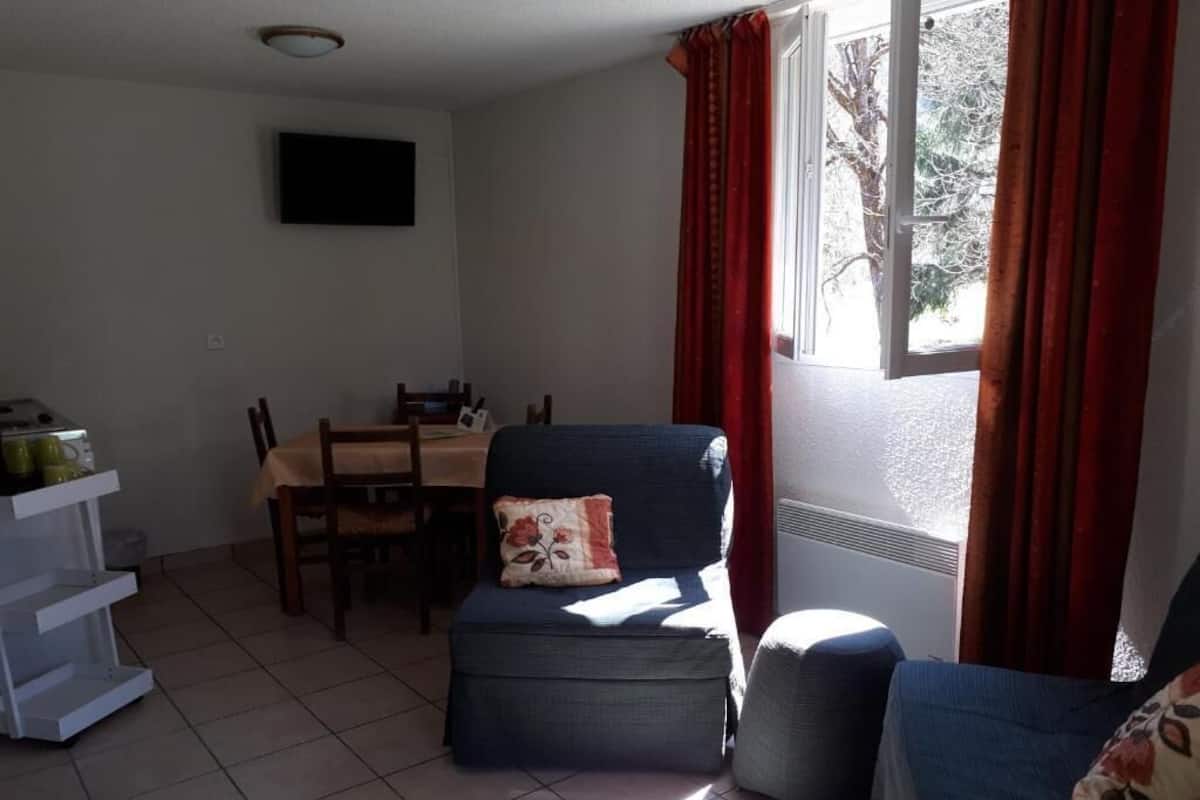 A room with a dining table, two chairs, a sofa, a TV mounted on the wall, and a window with curtains.
