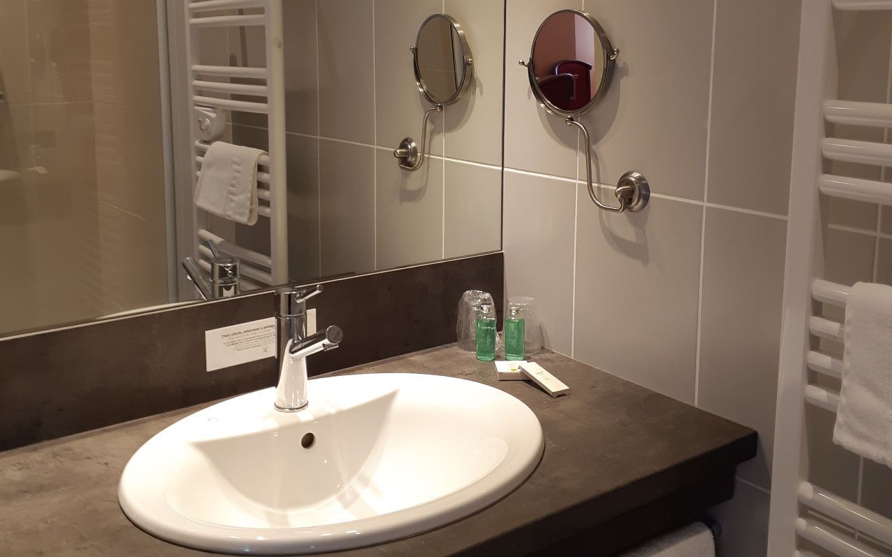 Ein Badezimmer mit einem weißen Waschbecken, zwei Spiegeln an der Wand, einem Handtuchhalter und einer Seifenspender.