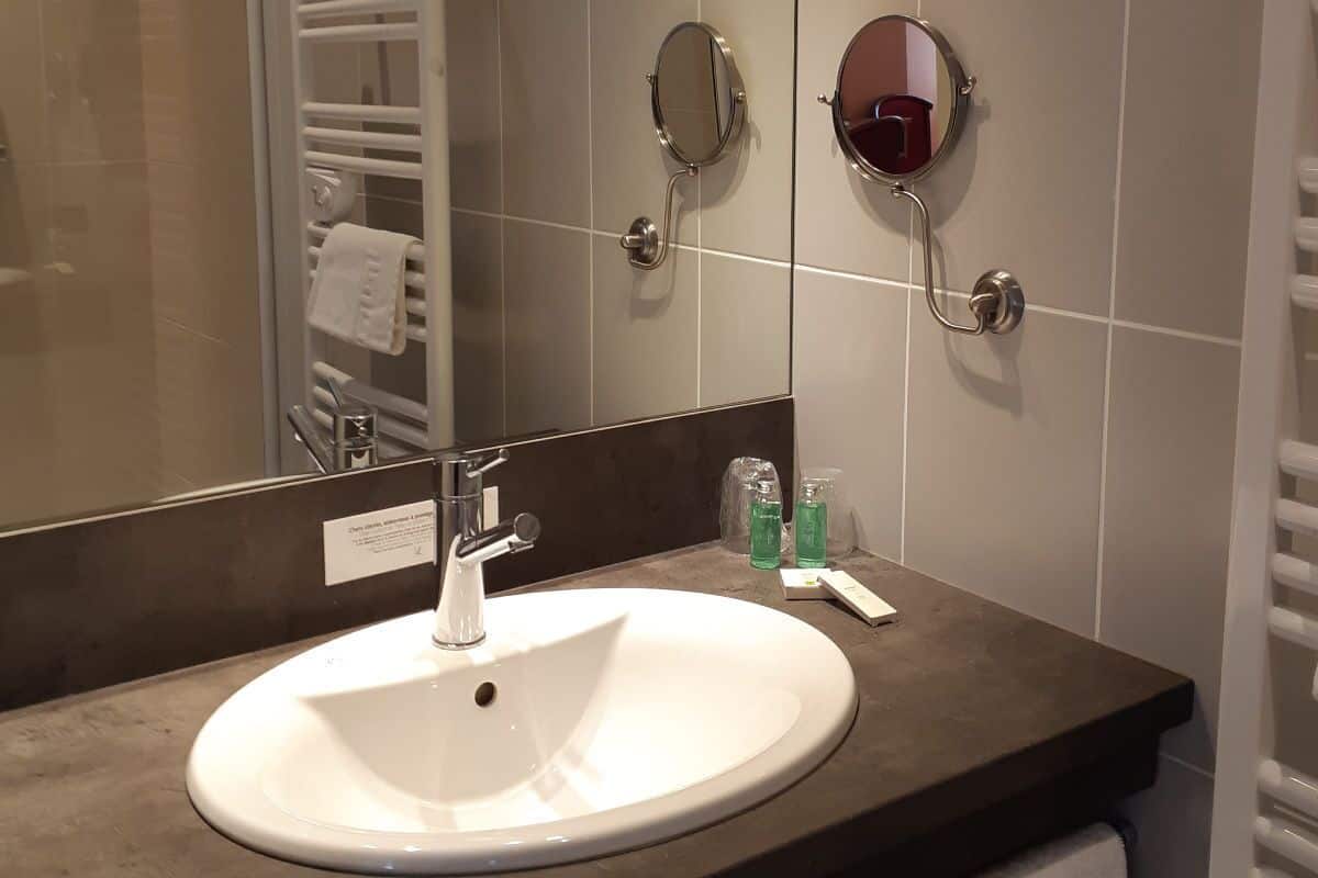 A bathroom with a white sink, two wall-mounted mirrors, a towel rack, and a soap dispenser.