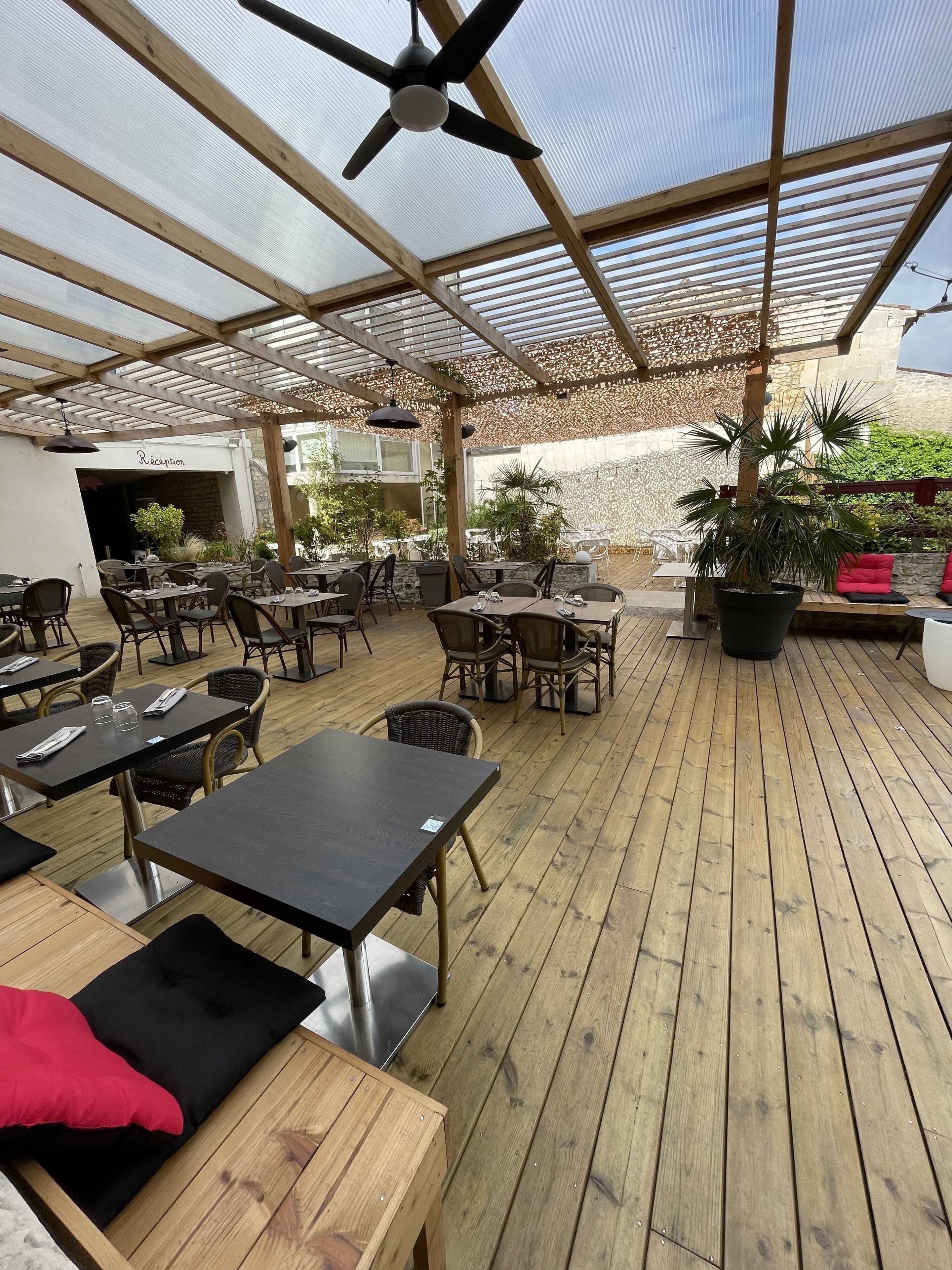 An outdoor patio with wooden tables and chairs, a ceiling fan, and a reception area.