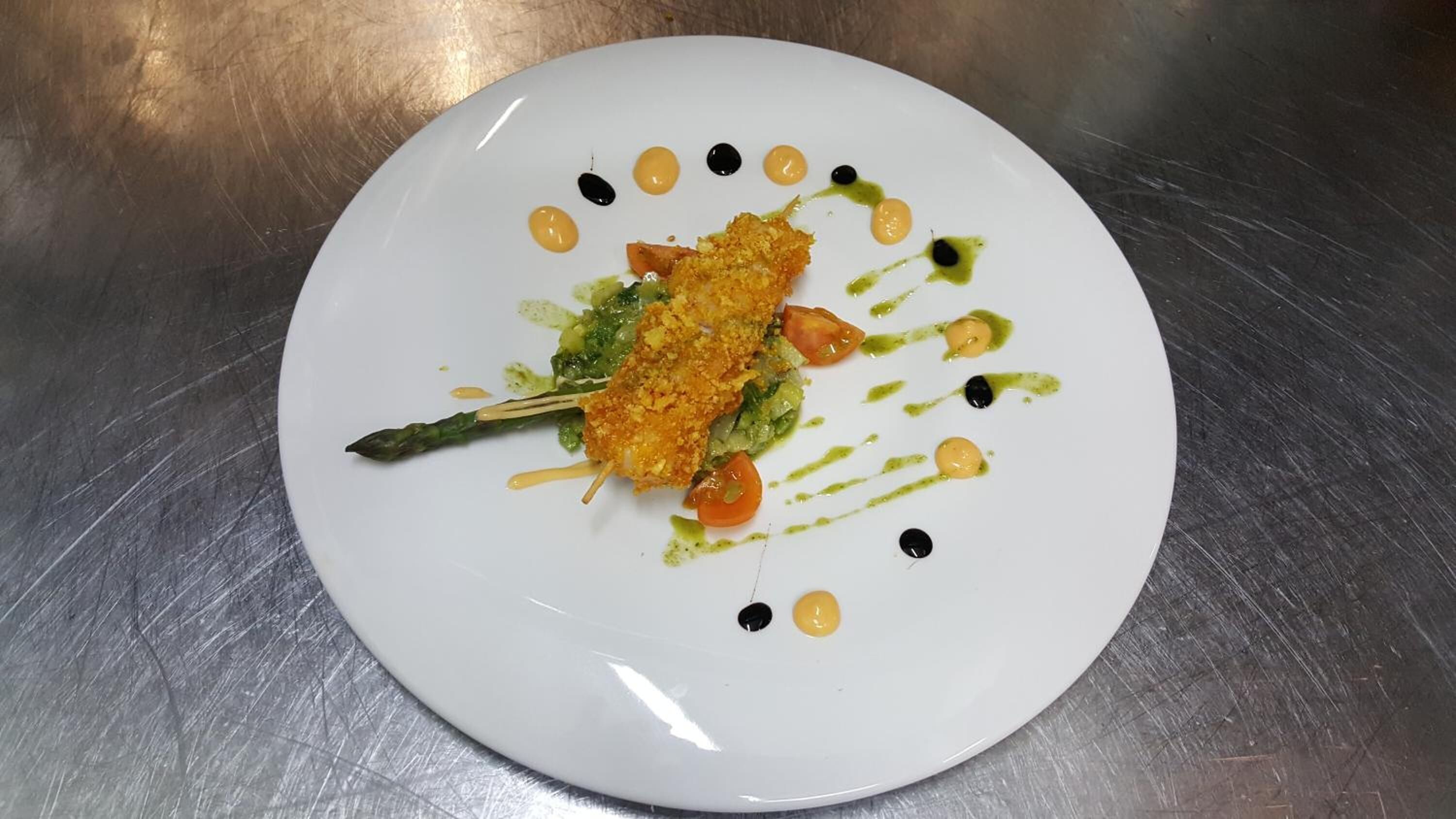 Ein weißer Teller mit einem Gericht aus paniertem Gemüse, Spargel und Tomaten, garniert mit grüner Soße und schwarzen Punkten.