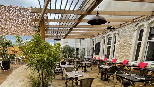 An outdoor patio with wooden tables and chairs, stone planters, and a pergola structure. - Hôtel de L'Ecu (Jonzac)