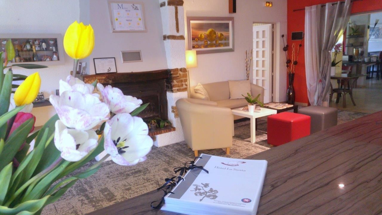 A hotel lobby with a fireplace, a sofa, a coffee table, and a red ottoman. There are flowers on the left and a brochure on the right.