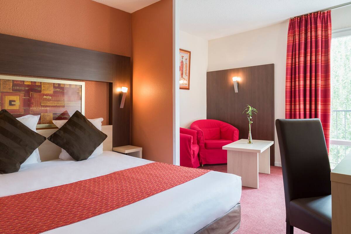 Una habitación de hotel con una cama grande, un sillón rojo, una mesita y una silla.