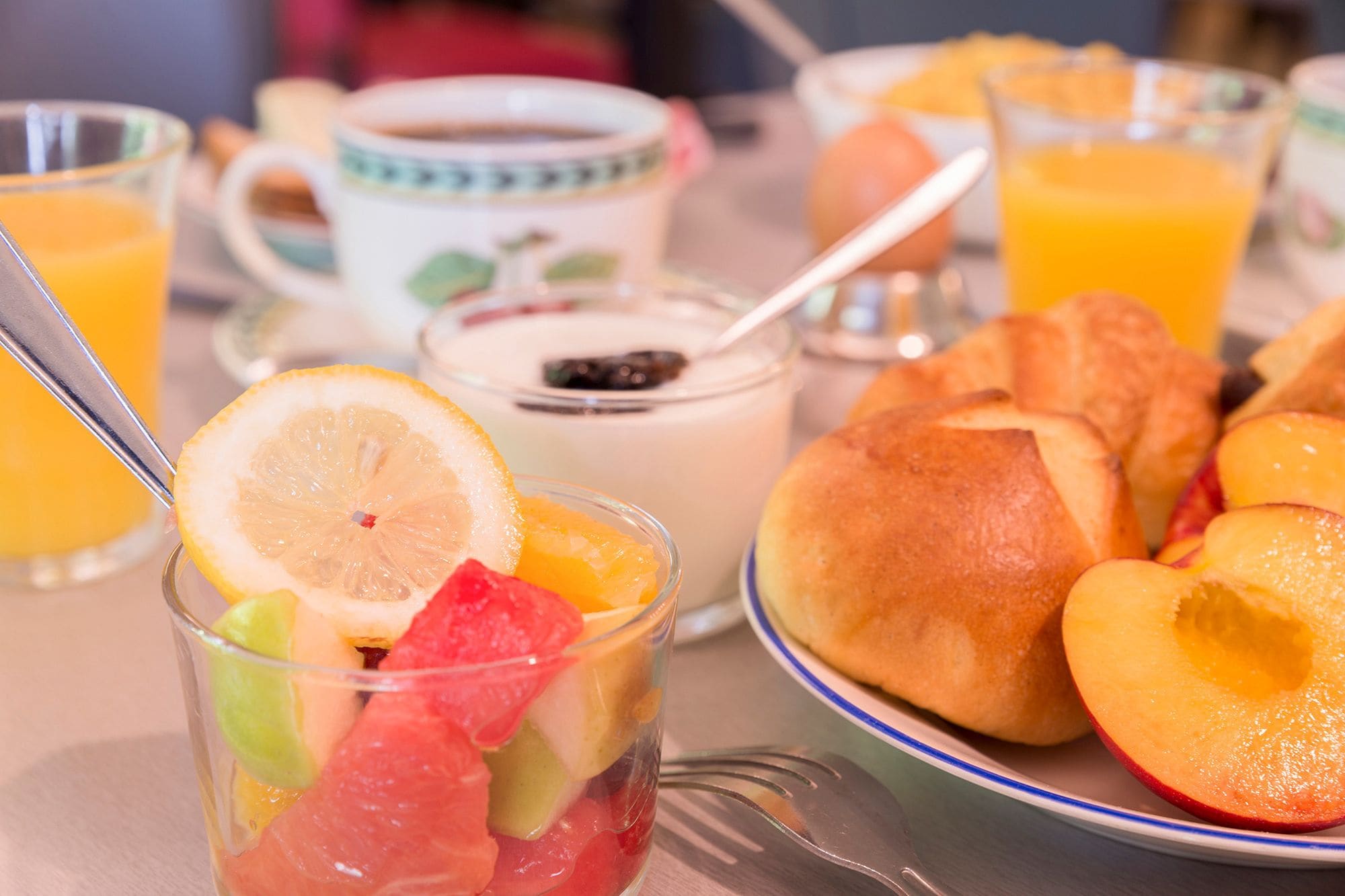 Una mesa de desayuno con un vaso de jugo de naranja, una taza de café, un tazón de yogur, un croissant y rodajas de melocotones.