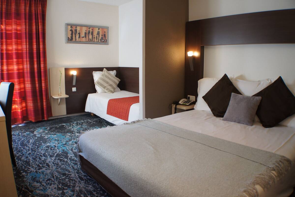 Habitación de hotel con dos camas, una cortina roja, un cuadro en la pared y una mesita de noche con un teléfono.