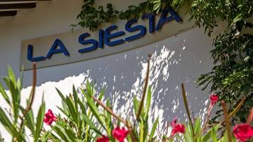 Valkoista seinää vasten on kyltti, jossa lukee ”LA SIERRA”. Kylttiä ympäröi vihreää lehtokasvillisuutta ja punaisia kukkia.
