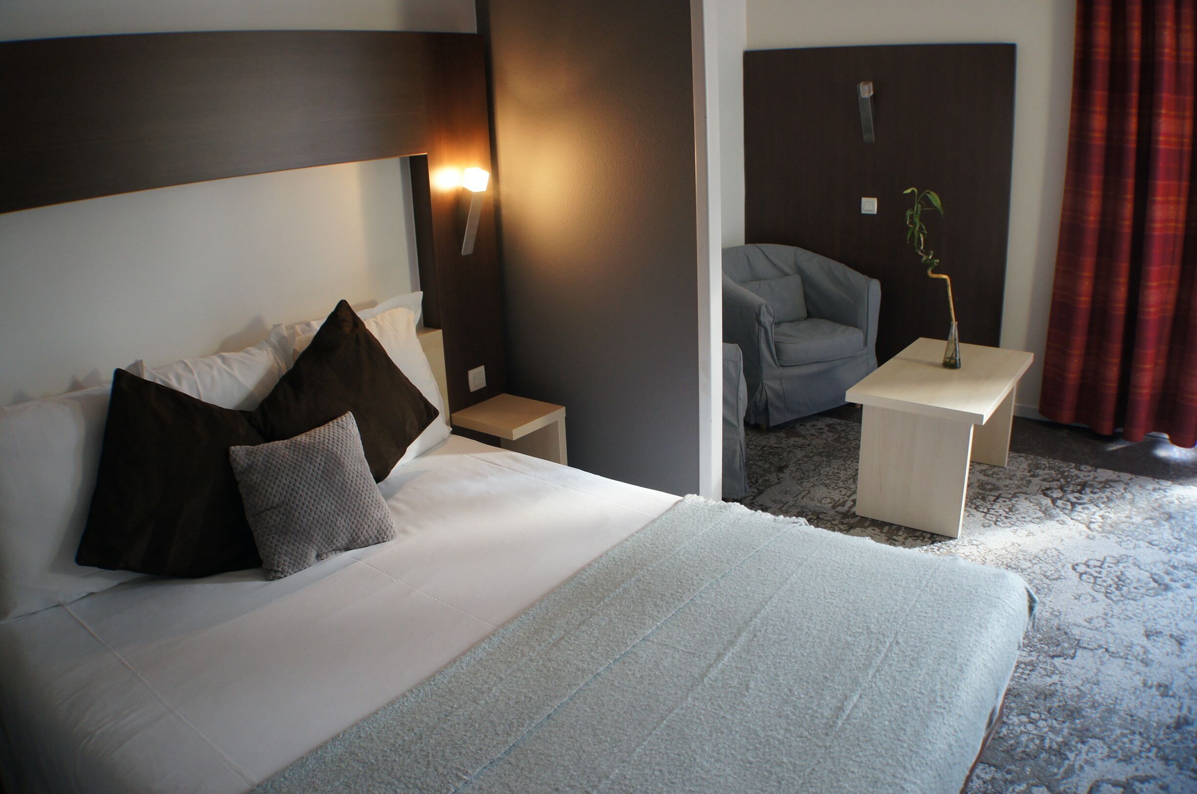 A hotel room with a large bed, a bedside table, a nightstand, a chair, and a small table with a plant.