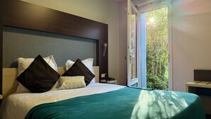 Habitación de hotel con cama, almohadas, mesita de noche con teléfono y ventana con vista a árboles.