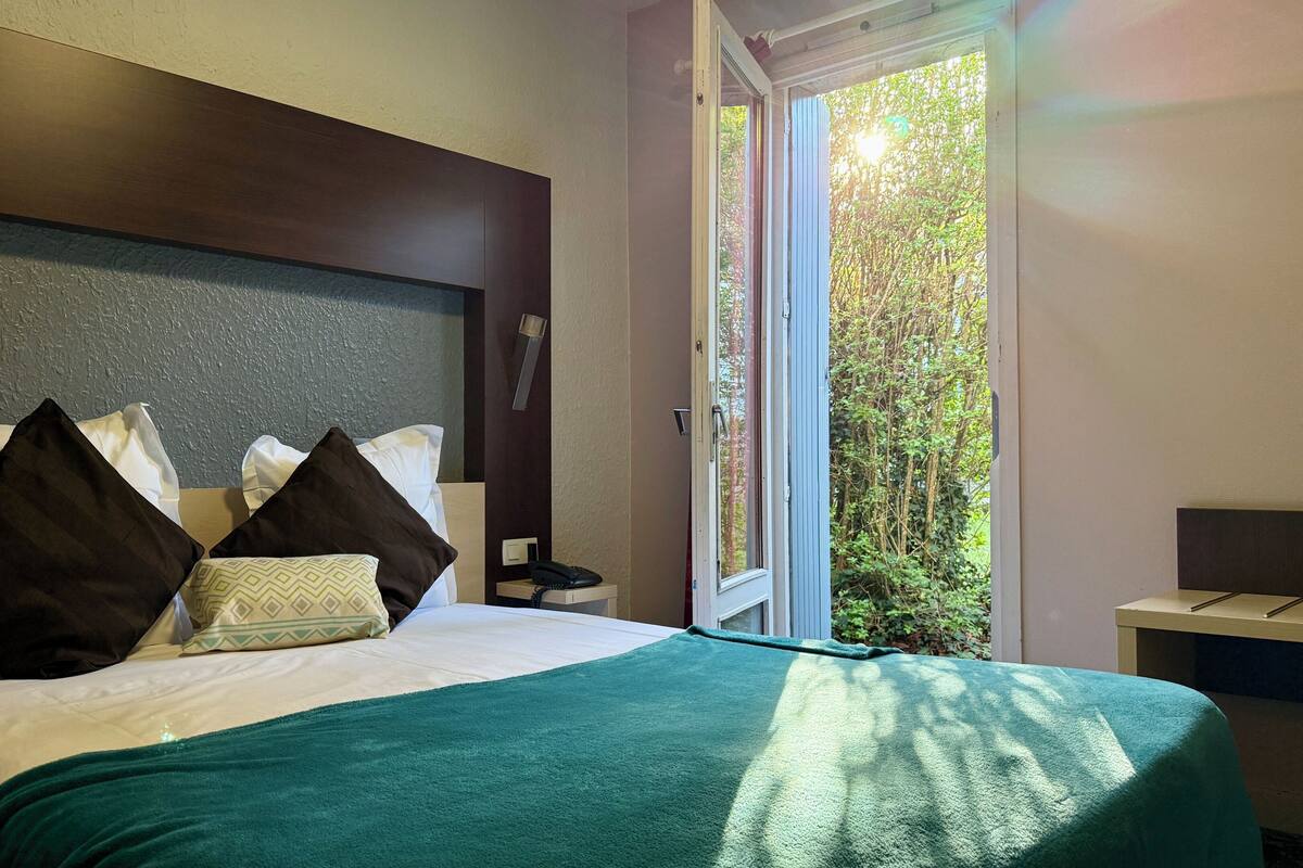 Habitación de hotel con cama, almohadas, mesita de noche con teléfono y ventana con vista a árboles.