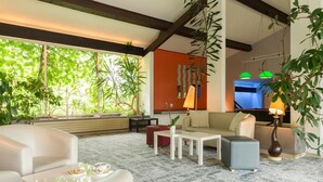 Una sala de estar moderna con un sofá, un sillón, una mesa de centro y un comedor con mesa y sillas. Amplias ventanas con vista a zonas verdes.