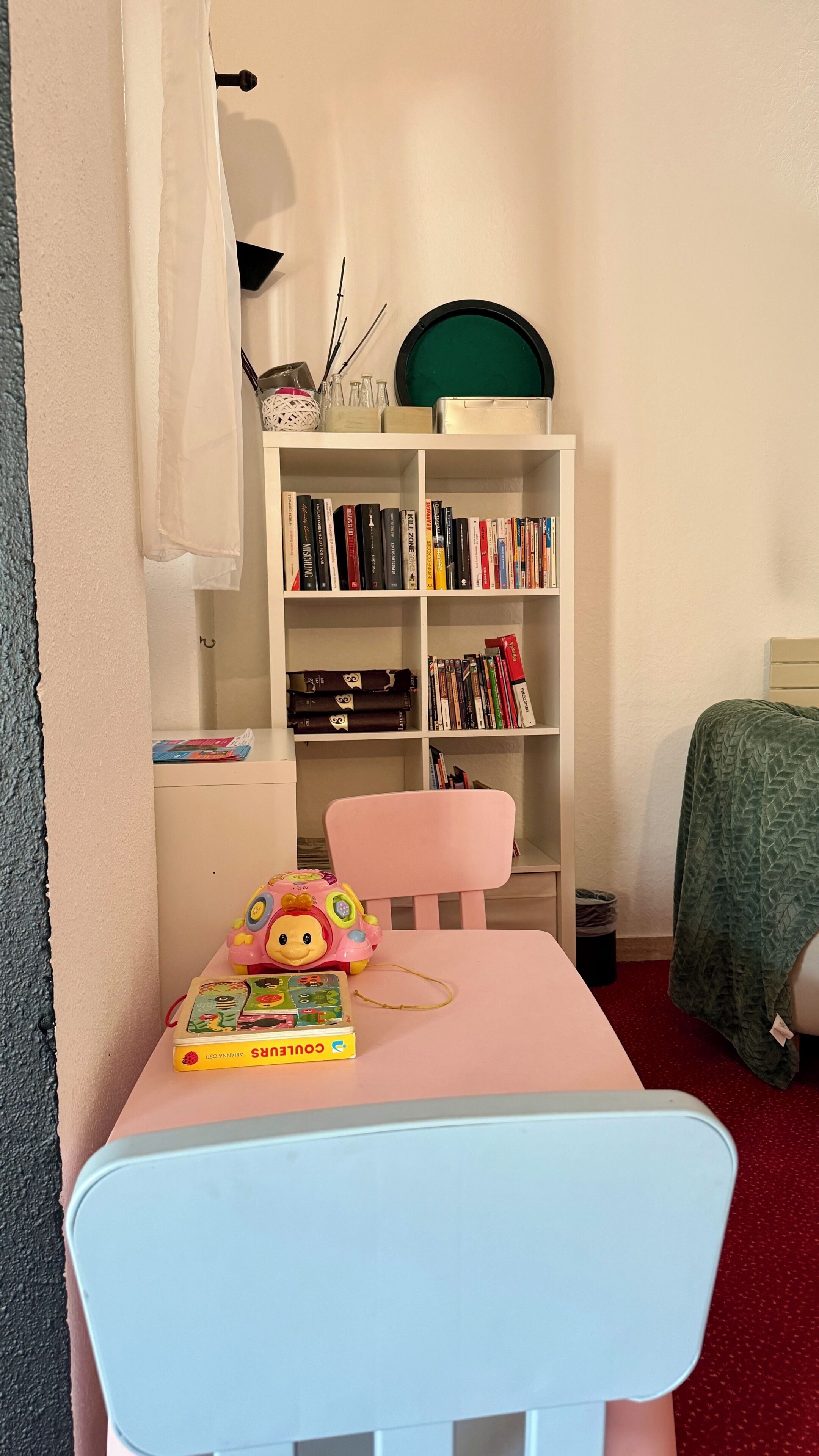A room with a white bookshelf, a pink chair, a small table with a toy and a book, and a bed with a green blanket.