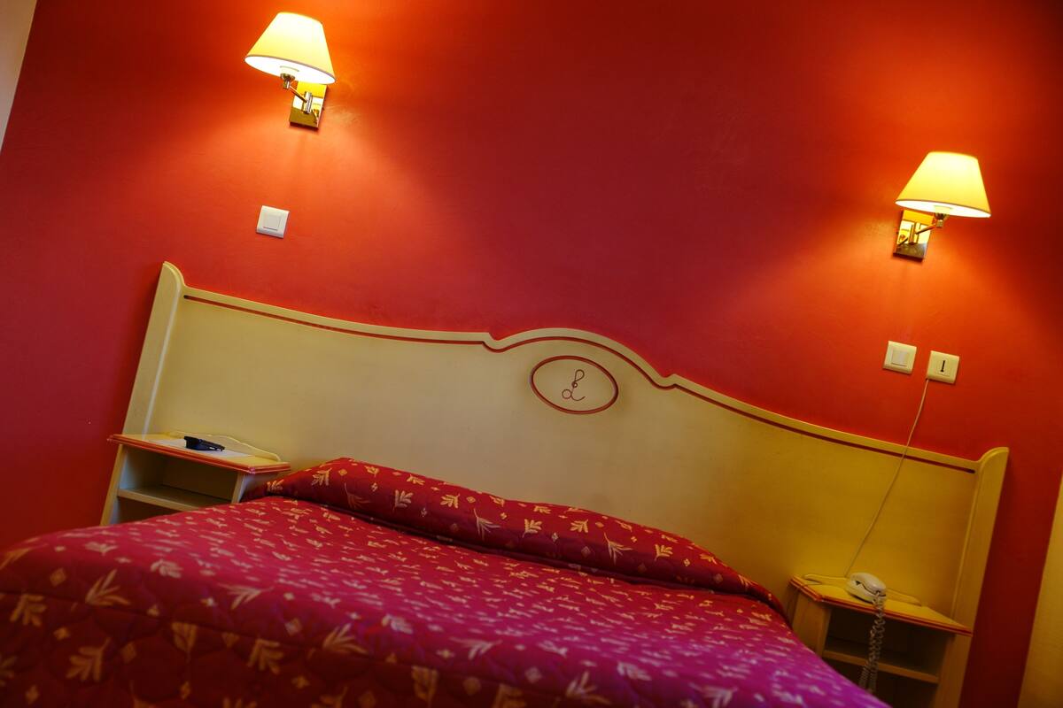 A hotel room with a red wall, two bedside lamps, a bed with a red and white patterned bedspread, and a nightstand with a telephone.