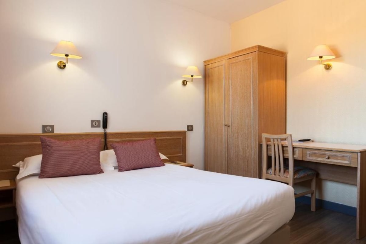 Une chambre d’hôtel avec un grand lit, une armoire en bois, un bureau avec une chaise et deux lampes fixées au mur.