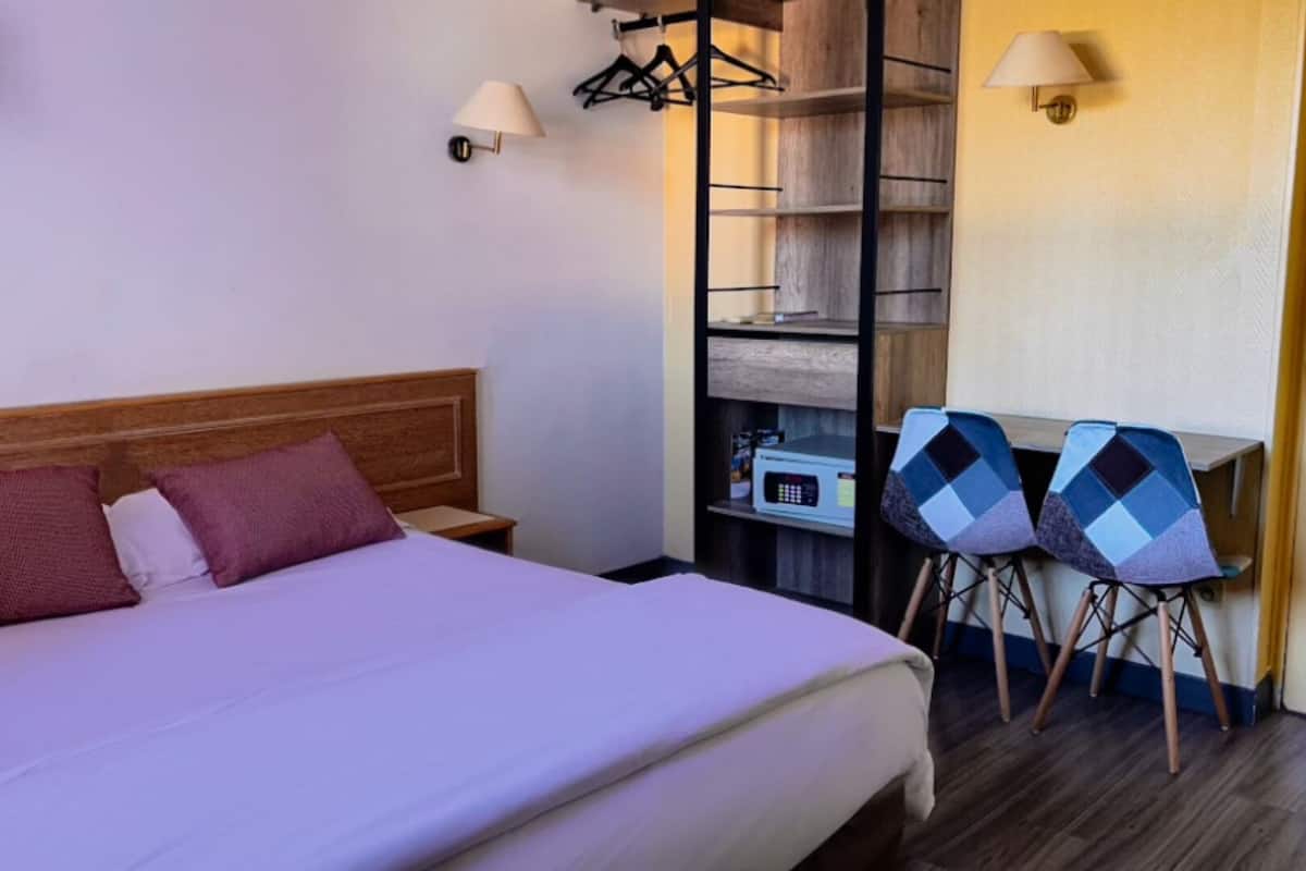 A hotel room with a bed, a desk with two chairs, a wardrobe, and a lamp.