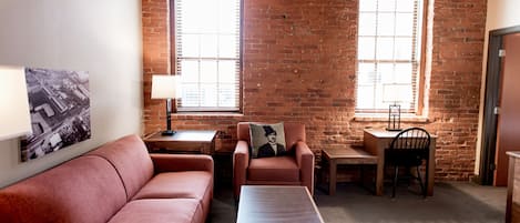 A cozy room with a brick wall, a red sofa, a wooden table, and two windows with blinds.
