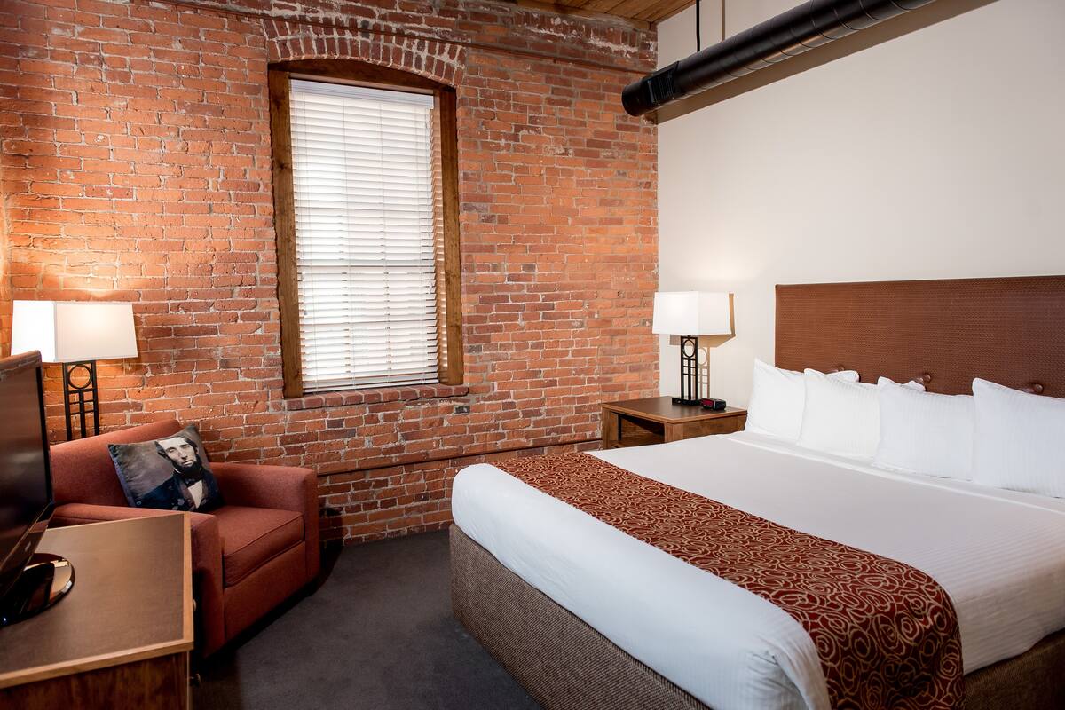 A hotel room with a brick wall, a large bed with white linens, a red sofa with a pillow, a nightstand with a lamp, and a window with blinds.