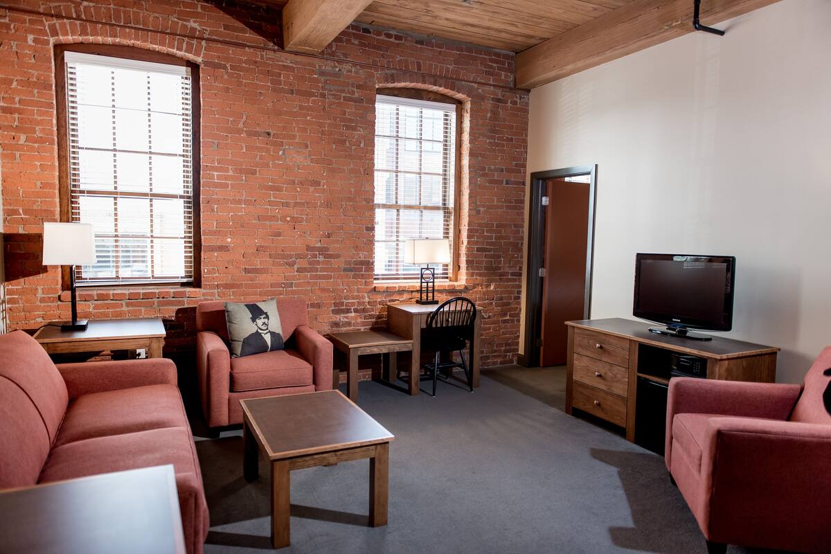 A furnished room with a brick wall, two windows with blinds, a television, a desk, a chair, and a sofa.