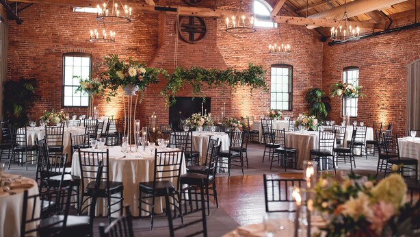 A spacious hall with brick walls, multiple tables set for dining, and elegant chandeliers.