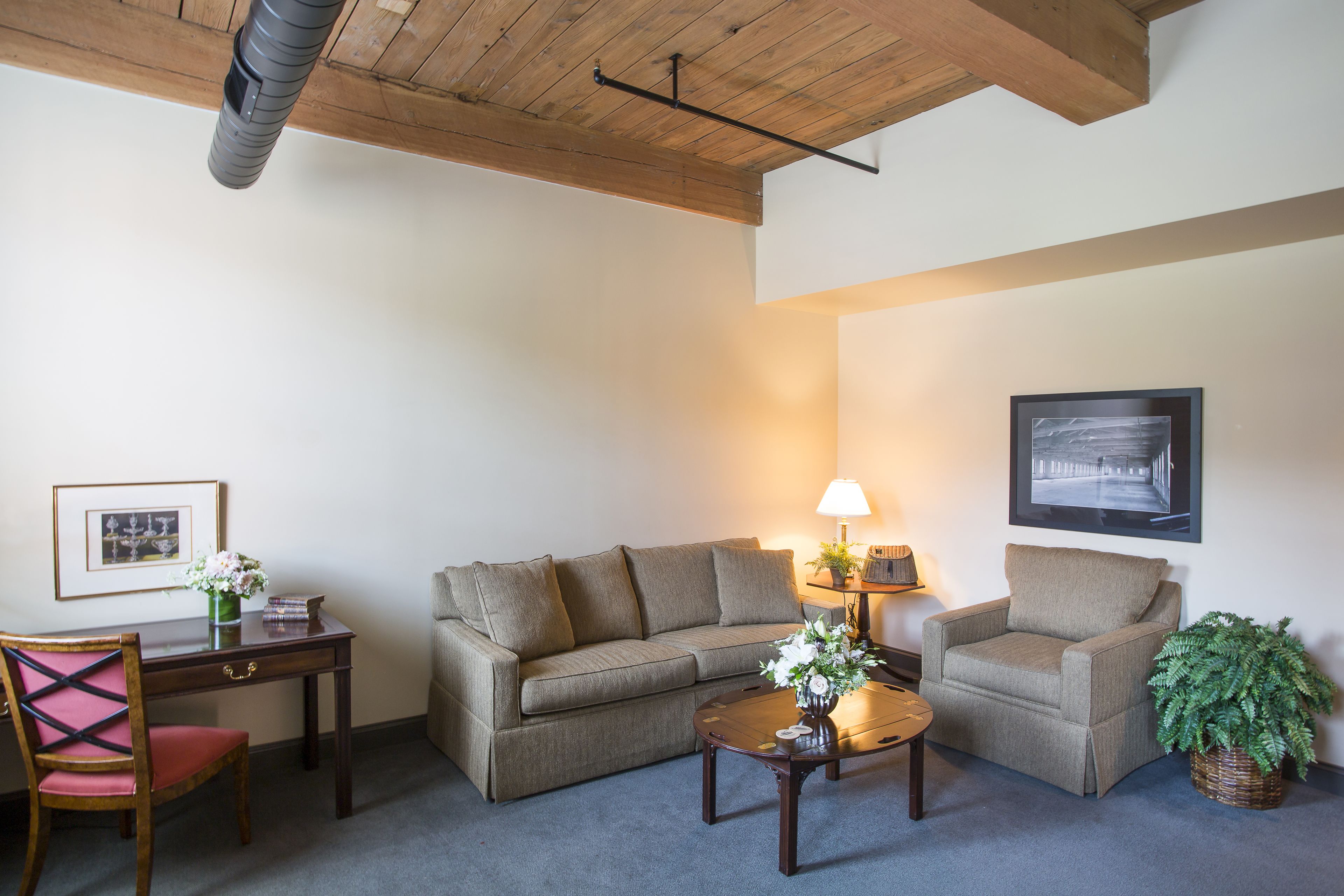 A living room with a wooden ceiling, a sofa, a chair, a small table with a lamp, and a framed picture on the wall.