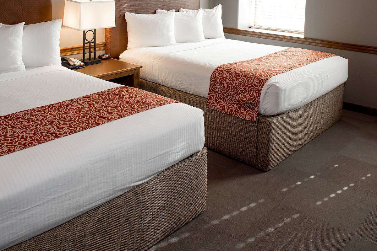 A hotel room with two beds, a wooden ceiling, a window, and a nightstand with a lamp.