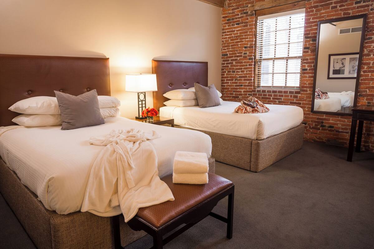 A hotel room with two beds, a bench, a nightstand with a lamp, and a large mirror.
