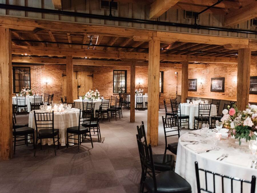 A spacious hall with multiple round tables, each set with white tablecloths and black chairs, featuring floral centerpieces.