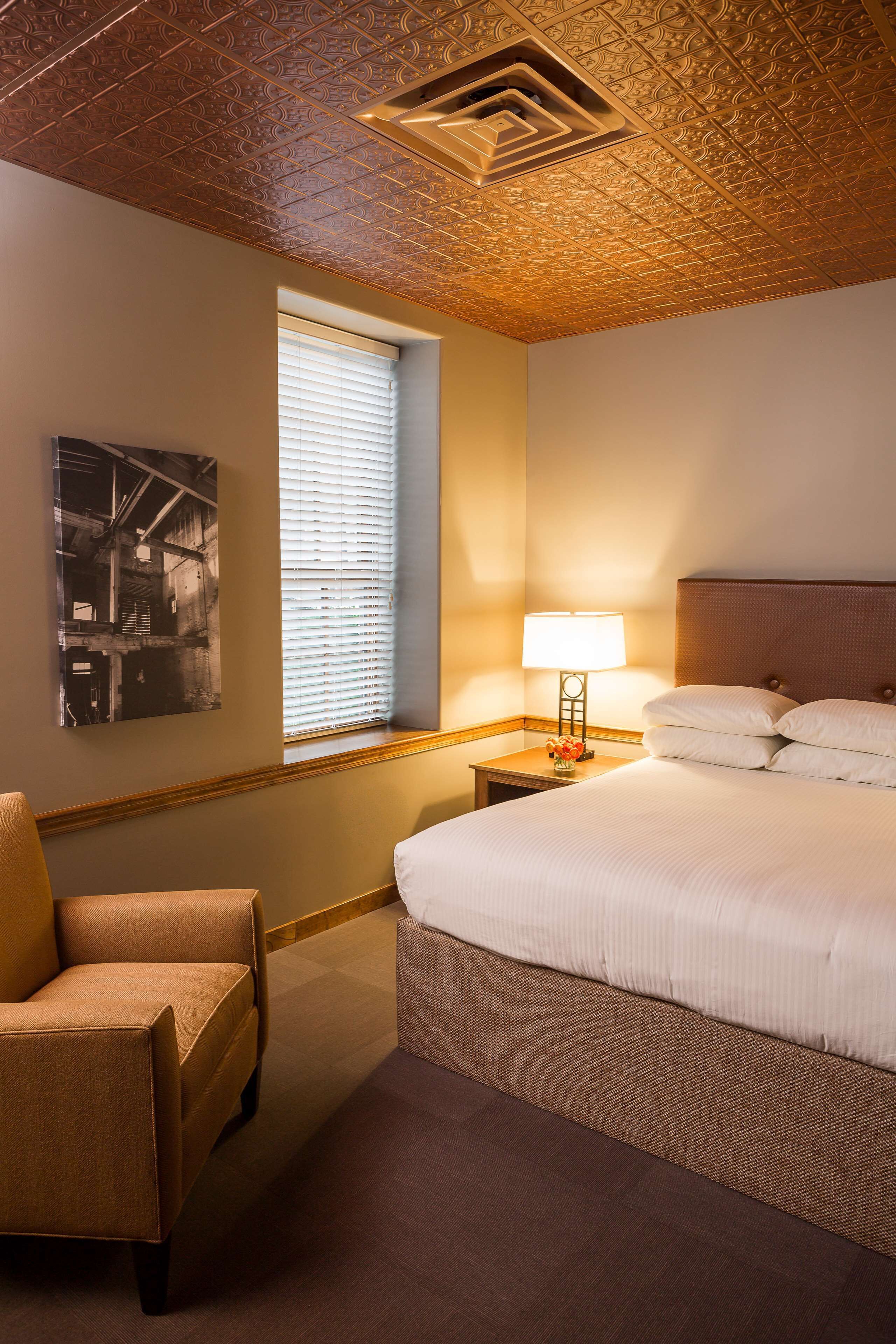 A hotel room with a large bed, a nightstand with a lamp, a chair, a window with blinds, and a ceiling with a decorative pattern.
